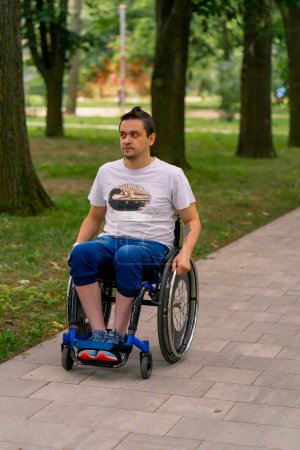 Foto de Inclusividad Un hombre con discapacidad monta una silla de ruedas a lo largo del camino del parque de la ciudad contra el fondo de los árboles - Imagen libre de derechos