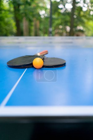 Foto de Dos raquetas de tenis y una pelota de tenis naranja se encuentran en una mesa de tenis azul junto a una red en un juego de ping pong en el parque de la ciudad - Imagen libre de derechos