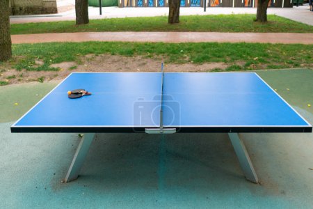 Photo for Two tennis rackets and an orange tennis ball lie on a blue tennis table next to a net in a city park ping pong game - Royalty Free Image