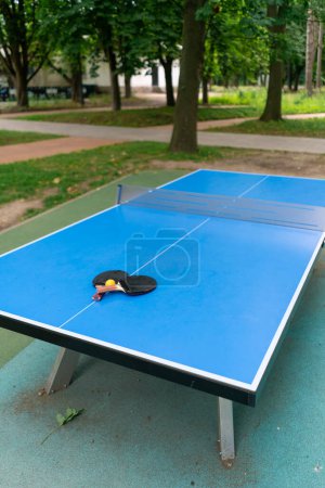 Photo for Two tennis rackets and an orange tennis ball lie on a blue tennis table next to a net in a city park ping pong game - Royalty Free Image