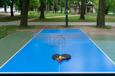 Téléchargez les photos : Deux raquettes de tennis et une balle de tennis orange reposent sur une table de tennis bleue à côté d'un filet dans un jeu de ping-pong dans un parc de la ville - en image libre de droit