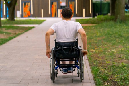 Foto de Inclusividad Un hombre con discapacidad monta una silla de ruedas a lo largo del camino del parque de la ciudad contra el fondo de los árboles Volver ver - Imagen libre de derechos