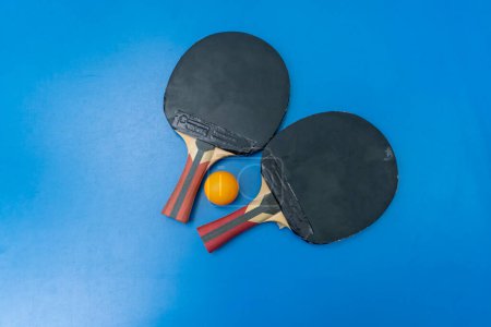 Foto de Dos raquetas de tenis y una pelota de tenis naranja se encuentran en una mesa de tenis azul junto a una red en un parque de la ciudad primer plano del juego de ping pong - Imagen libre de derechos