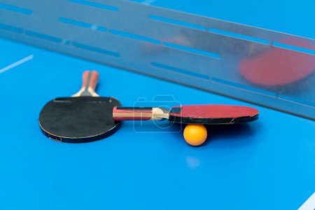 Photo for Two tennis rackets and an orange tennis ball lie on a blue tennis table next to a net in a city park close-up of ping pong game - Royalty Free Image