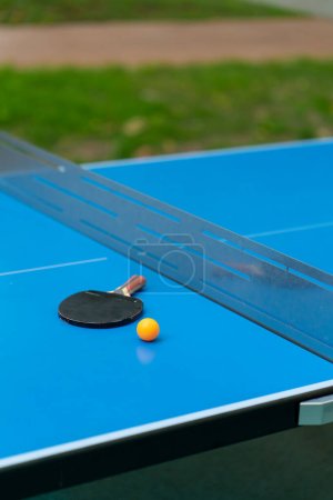 Foto de Una raqueta de tenis y una pelota de tenis naranja se encuentran en una mesa de tenis azul junto a una red en un parque de la ciudad primer plano del juego de ping pong - Imagen libre de derechos