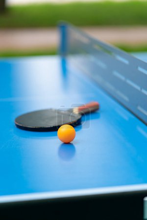 Photo for A tennis racket and an orange tennis ball lie on a blue tennis table next to a net in a city park close-up of ping pong game - Royalty Free Image