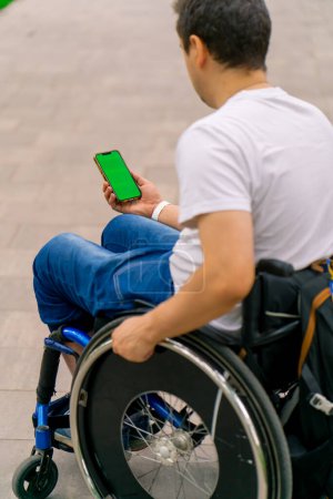 Foto de Inclusividad Un hombre con discapacidad en una silla de ruedas sostiene un teléfono con una pantalla verde abierta en la vista del parque de la ciudad desde atrás - Imagen libre de derechos
