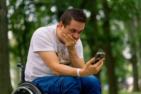 Photo for Inclusiveness A man with disabilities in a wheelchair looks into a smartphone and marvels at what he is holding in city park - Royalty Free Image