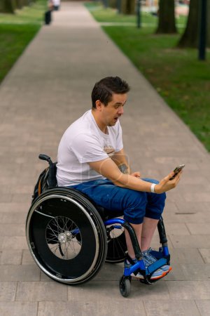 Foto de Inclusividad Un hombre con discapacidad en una silla de ruedas mira en un teléfono inteligente y se maravilla de lo que está sosteniendo en el parque de la ciudad - Imagen libre de derechos