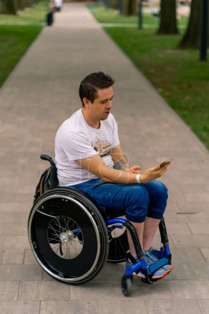 Téléchargez les photos : Inclusivité Un homme handicapé en fauteuil roulant regarde dans un smartphone et s'émerveille de ce qu'il tient dans le parc de la ville - en image libre de droit