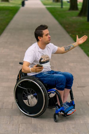 Téléchargez les photos : Inclusivité Un homme handicapé en fauteuil roulant tient un téléphone intelligent et montre de l'émotion dans un parc municipal sur fond d'arbres - en image libre de droit