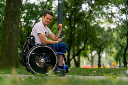 Photo for Inclusiveness Happy Man with disabilities in a wheelchair looking into a smartphone he holds in his hands in city park with trees in the background - Royalty Free Image