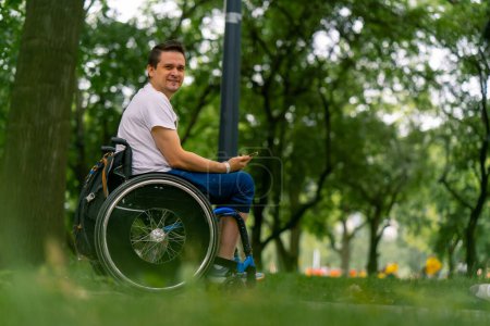 Foto de Inclusividad Happy Man con discapacidades en una silla de ruedas mirando a un smartphone que tiene en sus manos en el parque de la ciudad con árboles en el fondo - Imagen libre de derechos