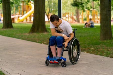 Foto de Inclusividad Retrato de un joven con discapacidad en silla de ruedas en un parque de la ciudad sobre el fondo de los árboles - Imagen libre de derechos