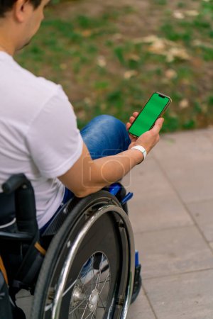 Photo for Inclusiveness A man with disabilities in a wheelchair holds a phone with an open green screen in city park view from behind - Royalty Free Image