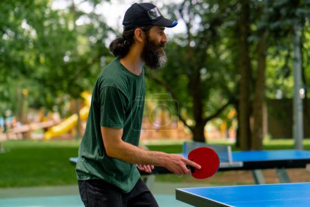 Foto de Un hombre mayor usando una gorra y sosteniendo una raqueta juega ping pong en un parque de la ciudad contra el telón de fondo de los árboles - Imagen libre de derechos
