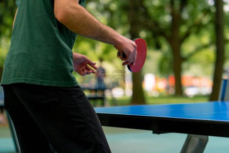 Foto de Un hombre mayor usando una gorra y sosteniendo una raqueta juega ping pong en un parque de la ciudad contra el telón de fondo de los árboles - Imagen libre de derechos