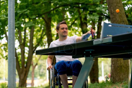 Foto de Inclusividad Un hombre discapacitado en silla de ruedas juega ping pong en un parque de la ciudad contra el telón de fondo de los árboles - Imagen libre de derechos