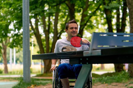 Téléchargez les photos : Inclusivité Un handicapé en fauteuil roulant joue au ping-pong dans un parc municipal sur fond d'arbres - en image libre de droit