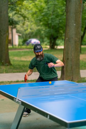 Foto de Inclusividad Un hombre anciano enfocado con el pelo largo y una barba gris juega ping pong en el parque de la ciudad - Imagen libre de derechos