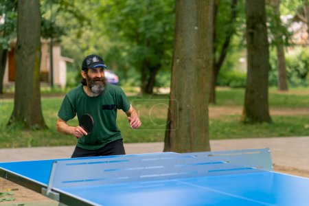 Photo for Inclusiveness An elderly focused man with long hair and a gray beard plays ping pong in city park - Royalty Free Image