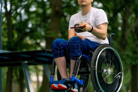 Foto de El hombre discapacitado inclusivo en una silla de ruedas junto a una mesa de pong de pin azul golpea una pelota naranja en una raqueta de tenis en el primer plano del parque de la ciudad - Imagen libre de derechos