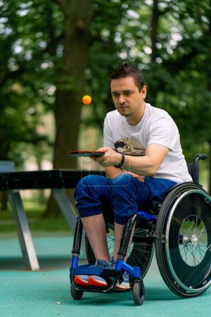 Téléchargez les photos : Un handicapé inclusif dans un fauteuil roulant à côté d'une table de pong à broches bleues frappe une balle orange sur une raquette de tennis dans un parc de la ville gros plan - en image libre de droit