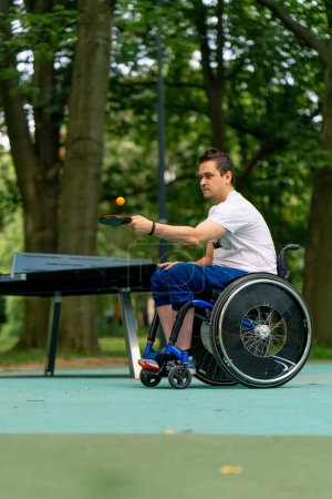 Photo for Inclusive  disabled man in a wheelchair next to a blue pin pong table hits an orange ball on a tennis racket in city park close-up - Royalty Free Image