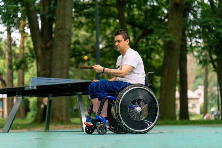 Foto de El hombre discapacitado inclusivo en una silla de ruedas junto a una mesa de pong de pin azul golpea una pelota naranja en una raqueta de tenis en el primer plano del parque de la ciudad - Imagen libre de derechos