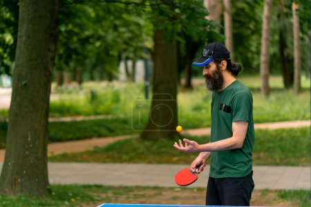 Photo for An elderly man next to a blue ping pong table hits an orange ball on tennis racket in a city park - Royalty Free Image