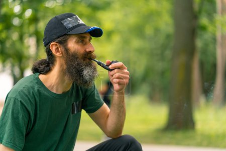 Téléchargez les photos : Un homme plus âgé avec les cheveux longs et la barbe grise fume pipe dans un parc municipal avec des arbres en arrière-plan - en image libre de droit