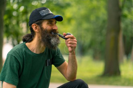 Téléchargez les photos : Un homme plus âgé avec les cheveux longs et la barbe grise fume pipe dans un parc municipal avec des arbres en arrière-plan - en image libre de droit