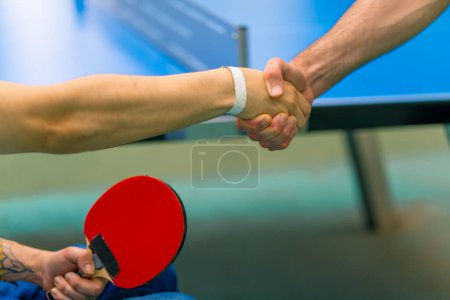 Téléchargez les photos : Un handicapé inclusif avec une raquette dans la main serre la main d'un homme plus âgé avant un match de ping-pong contre une table de tennis en arrière-plan - en image libre de droit