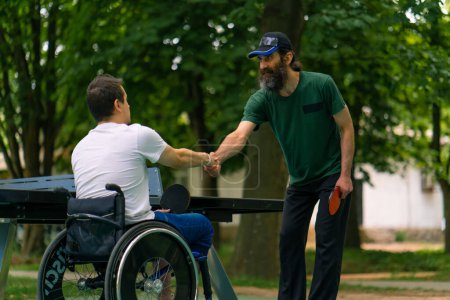 Photo for An inclusive disabled man with a racquet in his hand shakes hands with an older man before a game of ping pong against tennis table in the background - Royalty Free Image