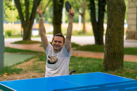 Photo for An inclusive disabled man with a racquet in his hand celebrates winning a game of ping pong against the backdrop of  city park - Royalty Free Image