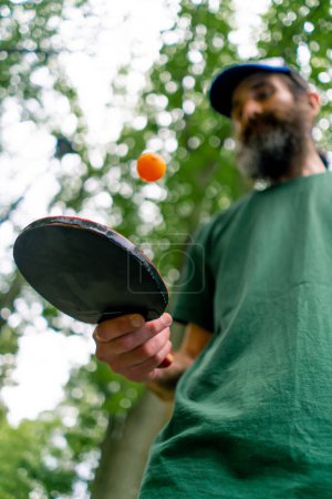 Foto de Un anciano junto a una mesa de ping pong azul golpea una pelota naranja en una raqueta de tenis en un parque de la ciudad - Imagen libre de derechos