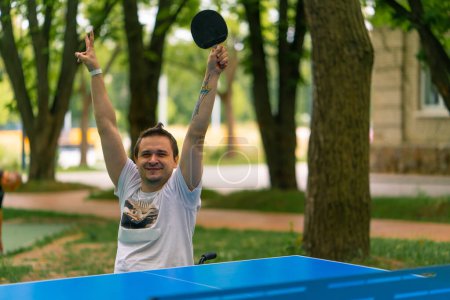 Foto de Un hombre discapacitado inclusivo con una raqueta en la mano celebra ganar un juego de ping pong en el contexto del parque de la ciudad - Imagen libre de derechos