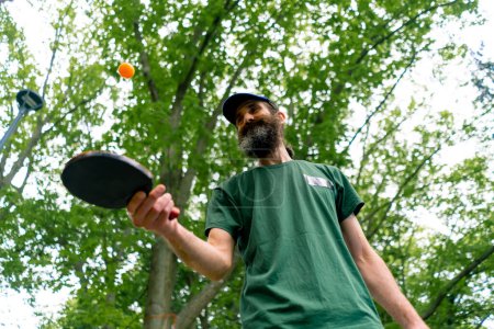 Téléchargez les photos : Un vieil homme près d'une table de ping-pong bleue frappe une balle orange sur une raquette de tennis dans un parc de la ville - en image libre de droit