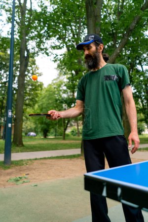 Téléchargez les photos : Un vieil homme près d'une table de ping-pong bleue frappe une balle orange sur une raquette de tennis dans un parc de la ville - en image libre de droit