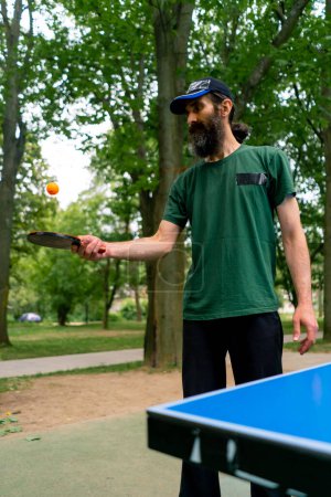 Foto de Un anciano junto a una mesa de ping pong azul golpea una pelota naranja en una raqueta de tenis en un parque de la ciudad - Imagen libre de derechos
