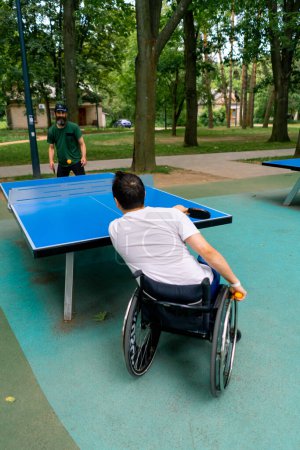 Foto de Inclusividad Un hombre discapacitado en silla de ruedas juega ping pong contra un anciano con barba gris en el parque de la ciudad - Imagen libre de derechos