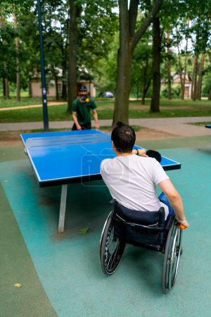Téléchargez les photos : Inclusivité Un handicapé en fauteuil roulant joue au ping-pong contre un vieil homme avec une barbe grise dans le parc de la ville - en image libre de droit