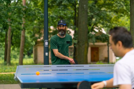 Foto de Inclusividad anciano jugando ping-pong contra un hombre con discapacidad que está en una silla de ruedas en el parque de la ciudad - Imagen libre de derechos