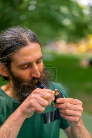 Téléchargez les photos : Portrait d'un homme âgé aux cheveux longs et à la barbe grise fumant une pipe dans un parc urbain sur fond d'arbres - en image libre de droit