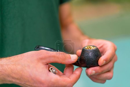 Téléchargez les photos : Un vieil homme en t-shirt vert fourre du tabac dans une pipe à fumer dans un parc de la ville sur fond d'arbres - en image libre de droit