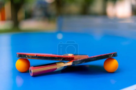 Téléchargez les photos : Deux raquettes de tennis et deux balles de tennis orange se trouvent sur une table de tennis bleue à côté d'un filet dans le parc de la ville jeu de ping-pong gros plan - en image libre de droit