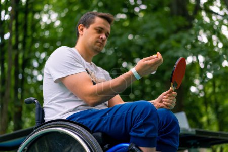 Foto de Inclusividad Un hombre discapacitado en una silla de ruedas con una raqueta y una pelota de ping-pong en sus manos cerca de una mesa de tenis en el parque - Imagen libre de derechos