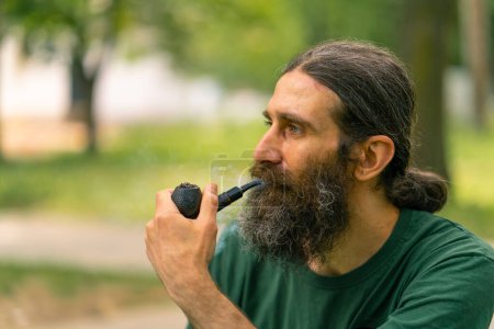 Téléchargez les photos : Portrait d'un homme âgé aux cheveux longs et à la barbe grise fumant une pipe dans un parc urbain sur fond d'arbres - en image libre de droit