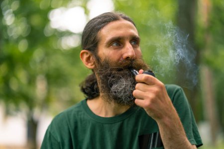 Foto de Retrato de un anciano de pelo largo y barba gris fumando una pipa en el parque de la ciudad sobre el fondo de los árboles - Imagen libre de derechos