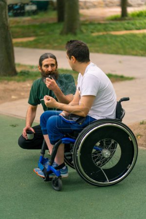Foto de Inclusividad Un hombre con discapacidad interactúa con un anciano fumando una pipa en un parque de la ciudad contra el telón de fondo de los árboles - Imagen libre de derechos